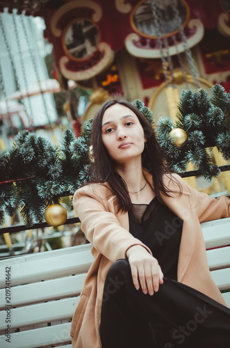 A beautiful young woman with long dark hair, wearing a black blouse, trousers, and a beige coat, sits on a park bench. Vertical photo.