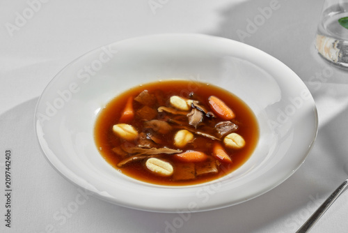 Gourmet mushroom broth soup with gnocchi and baby carrots on a white plate. Minimalist top view fine dining table setting with hard shadows.