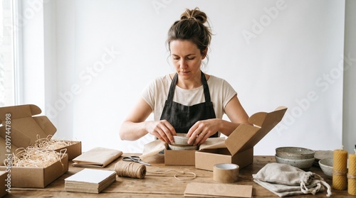 Small Business Owner Packing Orders: A focused woman entrepreneur meticulously packs handmade ceramic bowls into eco-friendly cardboard boxes, preparing shipments for her online store.
