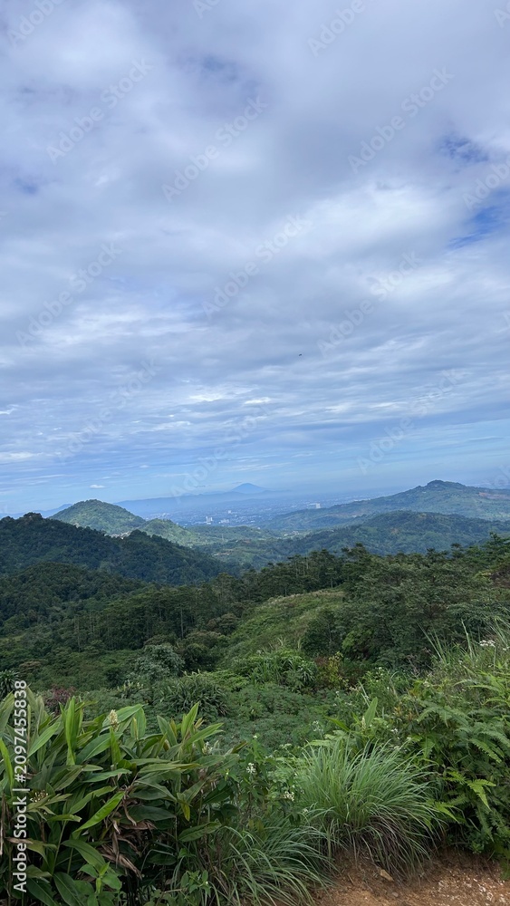 Fototapeta premium Vast Mountain Vista: A breathtaking panoramic view unfolds across a landscape of rolling, verdant mountains stretching towards a hazy horizon under a dramatic, cloud-filled sky.