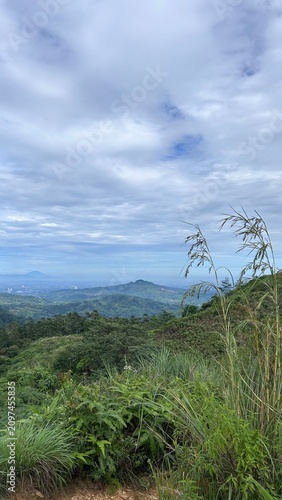 Obraz na plátne Verdant Hilltop Vista: A breathtaking panoramic view from a lush green hilltop overlooks rolling hills and distant mountains under a vast, cloud-dappled sky