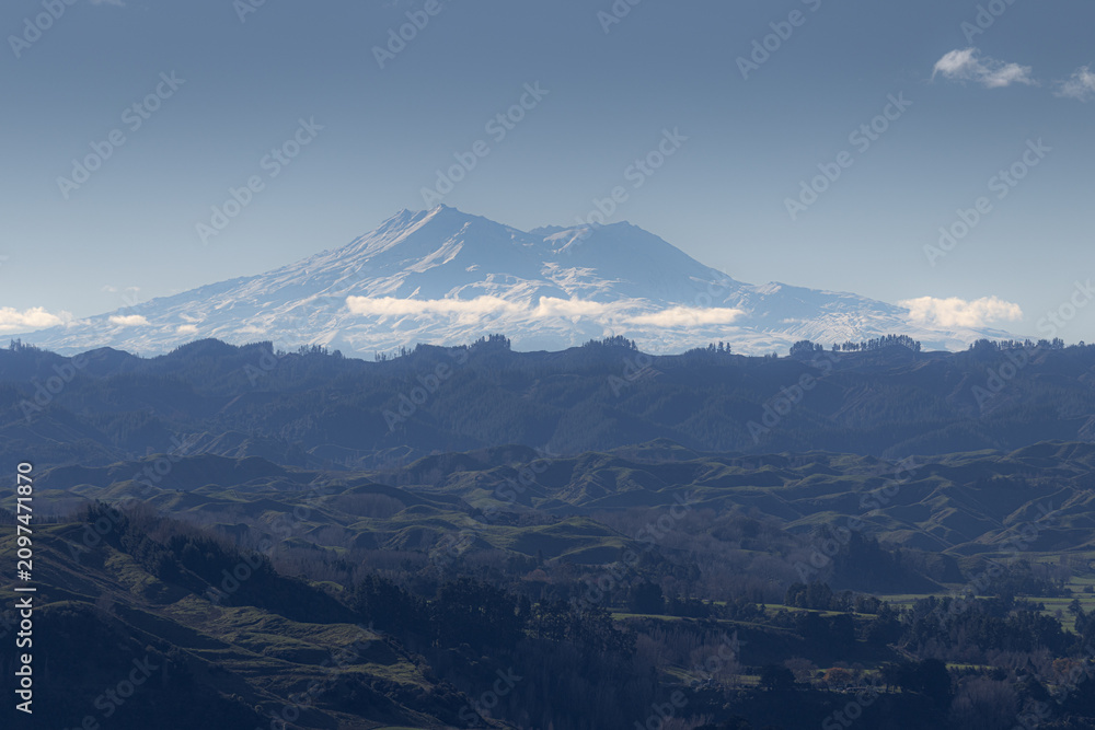 Fototapeta premium A majestic view of Mount Ruapehu, New Zealand, with snow-capped peaks under a clear blue sky. The foreground shows rolling hills covered in lush green vegetation.