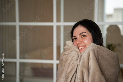Smiling young woman wrapped in a warm blanket indoors suggesting need for better heating. Winter home comfort and energy efficiency challenges