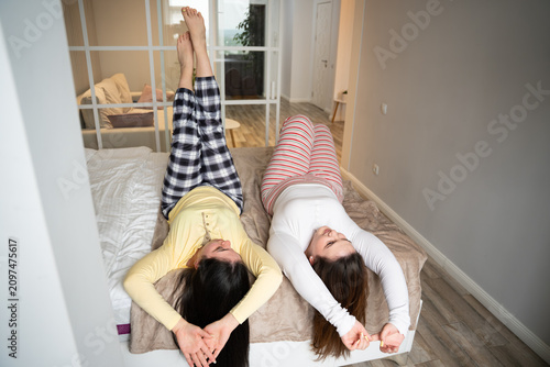 Two sisters lounging playfully on a bed indoors at home. Sibling bonding, family relationship, and playful home lifestyle