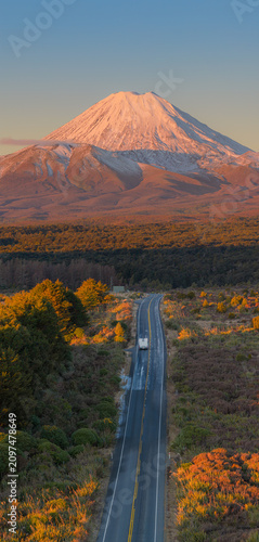 A scenic road winds towards Mt Ngauruhoe, a snow-capped volcano, in Tongariro National Park, New Zealand, during a golden sunset.