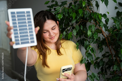 Young woman using smartphone while holding a portable solar panel indoors. Sustainable technology and off grid power innovation