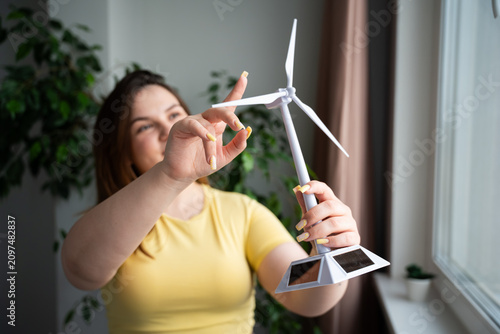 Young woman holding a small wind turbine model indoors near a window. Renewable energy education and green technology concept