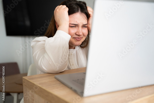 Stressed woman holding her head in front of a laptop at home office. Remote work burnout and deadline pressure in a digital workspace environment