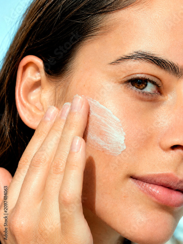 Close-up portrait of a young woman applying white skincare cream to her face. Vertical photo of a female spreading moisturizer on her cheek against a blue background. Daily facial hydration concept
