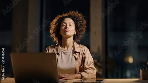 Young businesswoman practicing mindfulness meditation during a late night work break with laptop in a modern office for workplace mental wellbeing