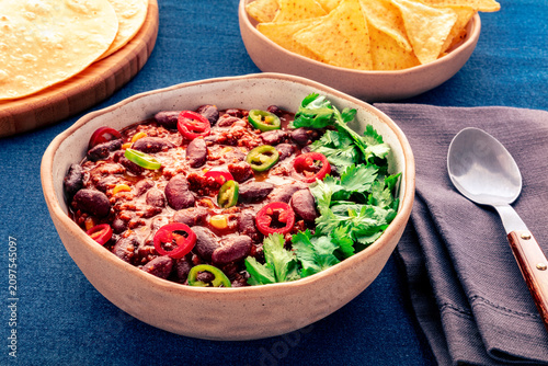 Spicy chili con carne, traditional Mexican beef stew, on blue background, with nachos and spoon