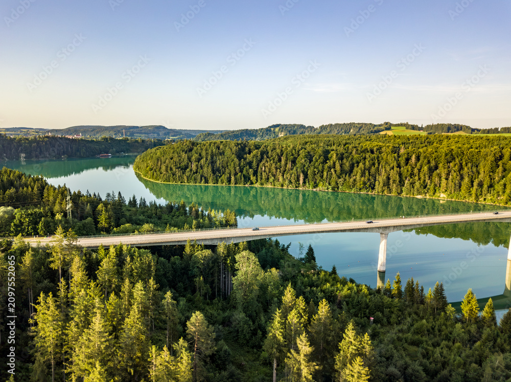 Obraz premium Aerial view of a long concrete viaduct bridge crossing the emerald green Lech river winding through dense forests in Bavaria, Germany.