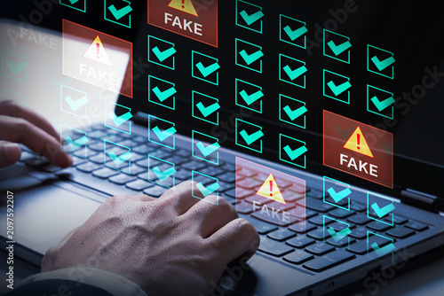 Digital Deception: Close-up on hands typing on a laptop, with a holographic interface displaying numerous checkboxes, some marked FAKE with warning signs.