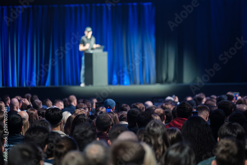 Large audience watching presenter on stage during technology conference. Blue curtains, spotlight, cameras, phones capturing keynote, focused crowd, professional atmosphere.