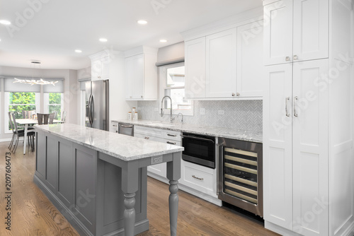 A modern kitchen with white cabinets, grey countertop, and a herringbone tile backsplash over a marble countertop looking towards a dining room.