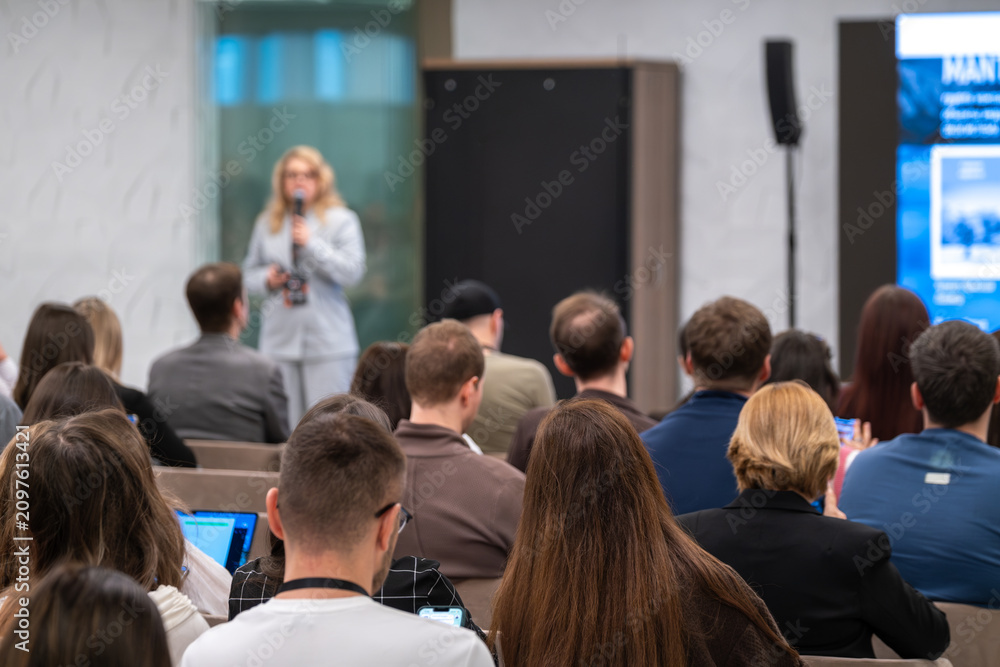 Fototapeta premium Business speaker presenting to attentive audience at indoor conference, professionals taking notes and using devices.