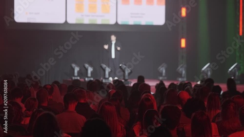 Business speaker presents on stage to large audience, projected slides visible, conference atmosphere, networking and learning mood.