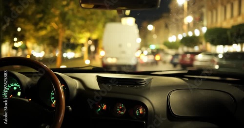 Car dashboard and steering wheel looking out at blurred night city traffic while driving behind a white van, POV