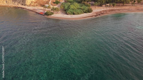 The video shows a beach with clear water and sandy shores. The view captures trees and buildings near the coastline. The sun is setting, casting light over the area.