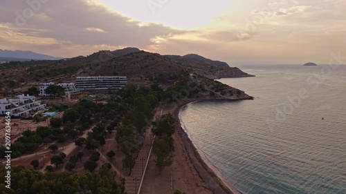 The camera captures a beach during sunset. The sun sets behind the hills, creating a warm glow over the water. People walk along the shore while others relax.
