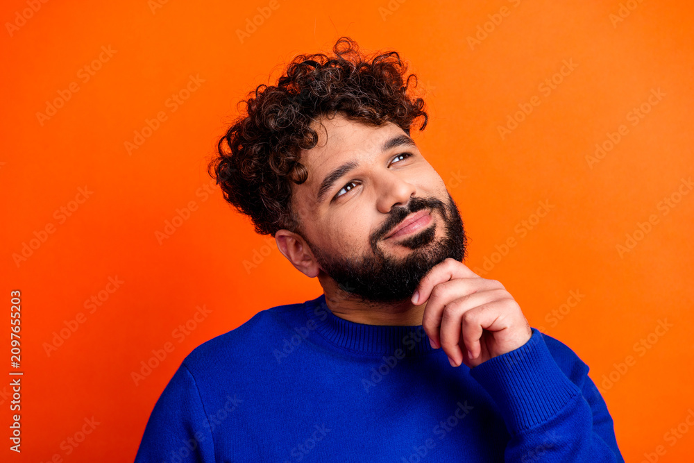 Obraz premium Thoughtful young man in blue sweater looking up with a smile on orange background, portrait of a bearded curly-haired person pondering an idea and feeling optimistic