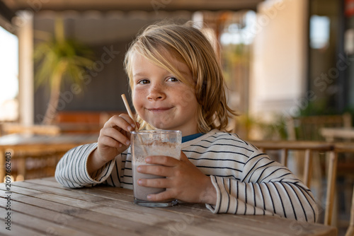 Smiling blond boy with freckles drinking chocolate milk at cafe table, holding glass with straw, cheerful childhood moment, cozy atmosphere, natural light, lifestyle scene