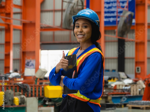 confident female African American engineer wearing blue hardhat and safety vest smiling while giving thumbs-up gesture inside modern manufacturing factory warehouse.