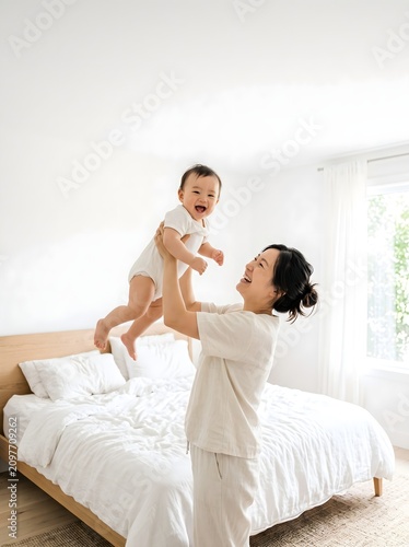 Joyful Asian Mother Lifting Happy Baby in Bright Minimalist Bedroom at Home