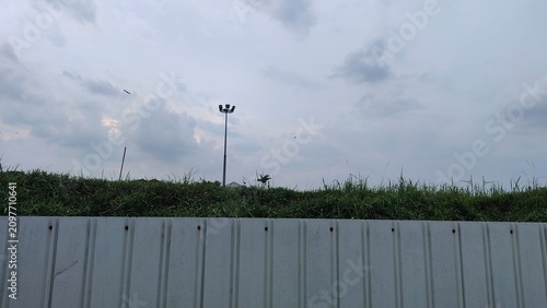 Wide Urban Industrial Background with Metal Fence Green Grass and Tall Light Pole under Overcast Sky Hero Header for Construction Site and Outdoor Events
