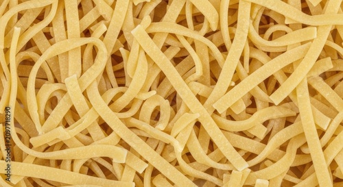 A pile of fresh, uncooked fettuccine pasta lies on a dark surface. The pasta strands are a light golden color and are arranged in a tangled, haphazard manner.