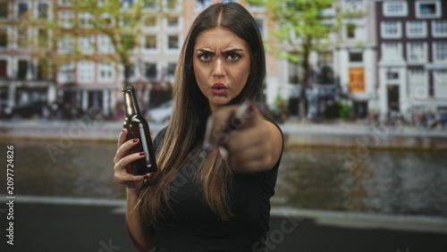 Foto Young hispanic woman holding beer bottle points finger at camera on a street by a canal with row houses and stern gaze; defiance accusation
