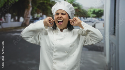 Canvastavla Young hispanic woman chef in white uniform and tall hat plugs ears with fingers, grimacing on a residential street with parked cars; annoyance stress tension