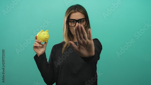 Foto Woman holding yellow piggy bank with palm out stop gesture in studio with teal backdrop; financial caution