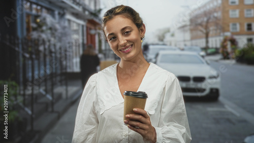 Fotografie Woman holding a takeaway coffee cup on a city street smiling gently in a white blouse; morning routine contentment