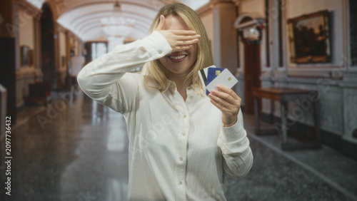 Woman holding creditcards and smiling while sometimes covering eyes with her hand in an ornate building hallway; joy financial confidence.