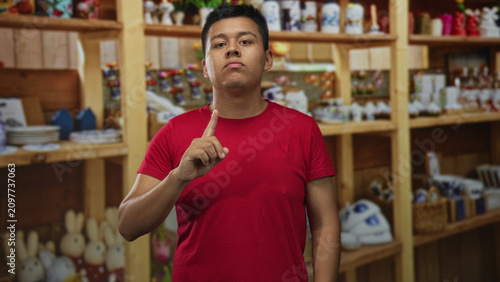 Canvastavla Man points finger upward in home decor shop aisle beside wooden shelves of ornaments while wearing red shirt; confidence