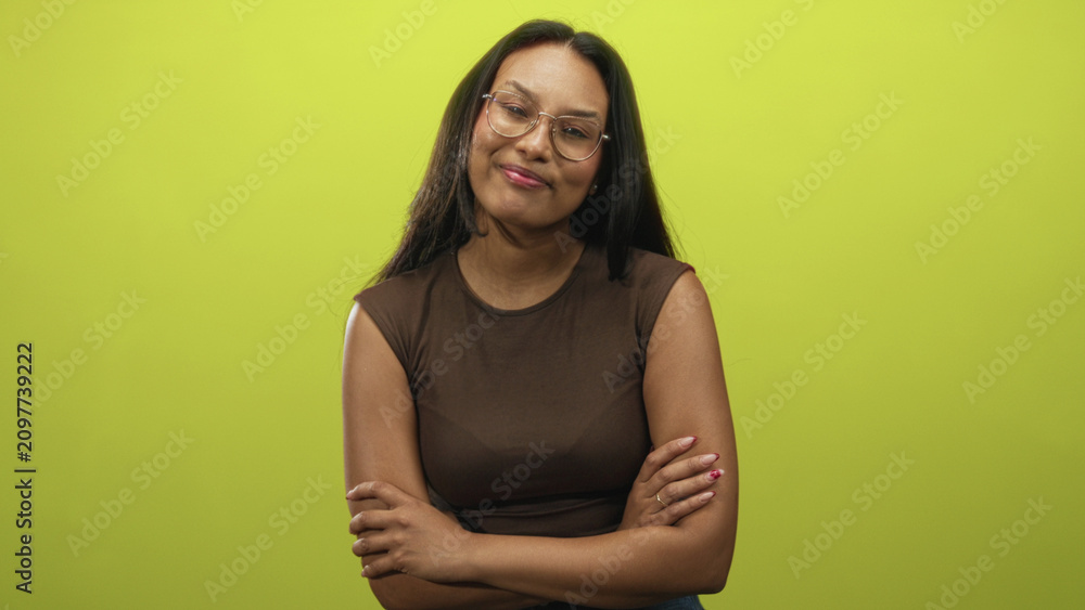 Obraz premium Woman, young brunette wearing glasses and brown sleeveless top, smiles and gives thumbs up while her arms are folded in studio; approval confidence.