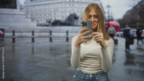 Foto Woman holding smartphone, pointing finger to screen and frowning on rainy street with red bus and lion statue; frustration tech problem
