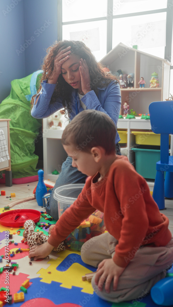 Fototapeta premium Teacher hand on forehead beside boy playing with plastic blocks and plush giraffe on foam mat in a building; tiredness patience.