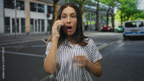 Foto Young woman holds smartphone to ear on a city street, glancing up and gesturing with her free hand while standing near parked cars; surprise concern