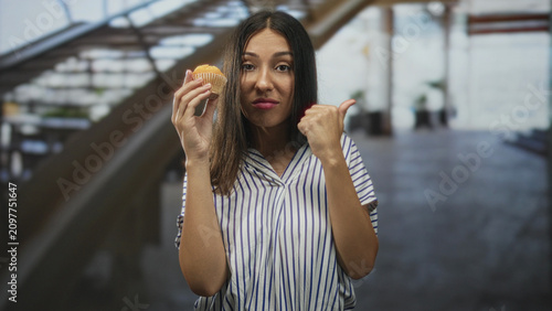 Konstfotografi Hispanic brunette woman holding a muffin in one hand while thumb pointing to the side with the other, standing near an escalator in a building; skepticism reluctance