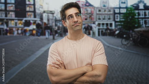 Fotografía Man with folded arms and visible forearms wearing pale pink t shirt and glasses looking upward on a cobblestone street; thoughtful contemplation