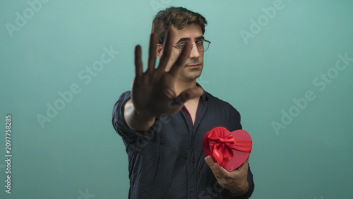 Bild auf Leinwand Man holding red heart shaped gift box with bow while extending palm out stop gesture in studio teal backdrop; rejection longing