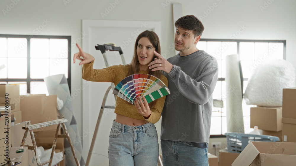 Naklejka premium Young woman and man holding a paint fan and pointing at color swatches in apartment living area; home renovation hopeful.