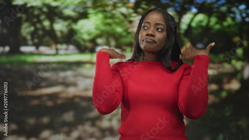 Foto Woman in red sweater raises hands in shrug gesture with palms visible against a park backdrop in a studio set; annoyance resignation