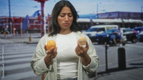 Fotografie Woman holding an orange in her left hand and a muffin in her right, eyes closed and frowning while comparing choices on a busy street; indecision temptation choice