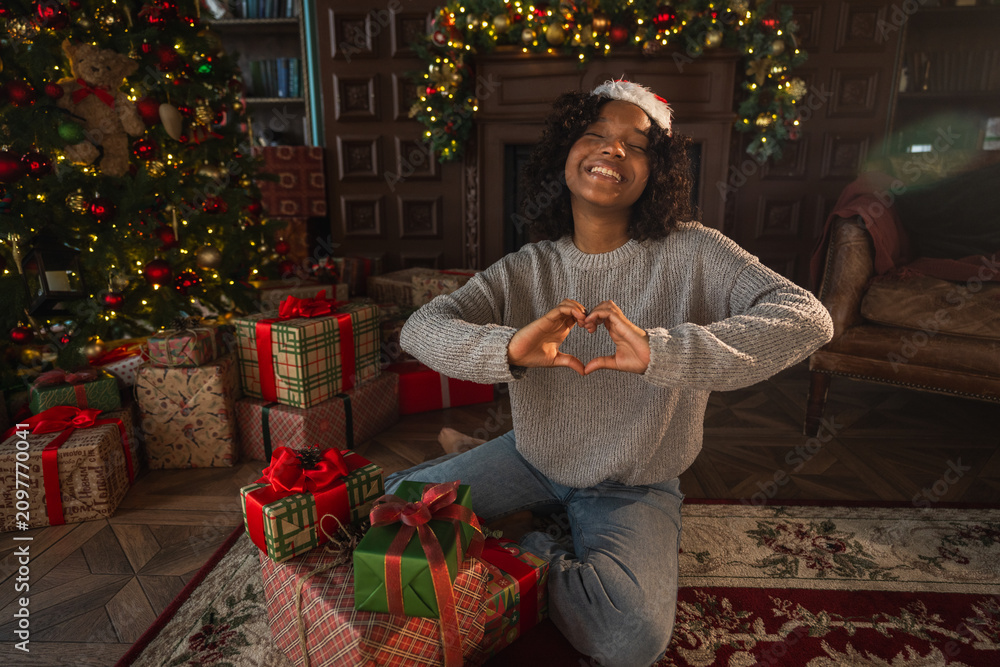Obraz premium Merry Christmas. African American woman smiling making heart shape with hands near Christmas tree. Happy girl in Santa hat in living room with fireplace Christmas gift boxes. Christmas eve at home