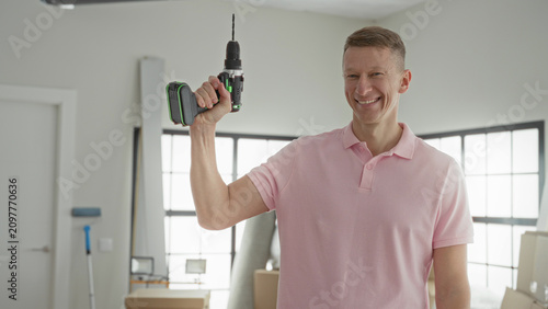 Billede på lærred Man in pink polo holds a cordless drill aloft with raised hand and visible forearm inside a bright residential building room; confidence home improvement progress