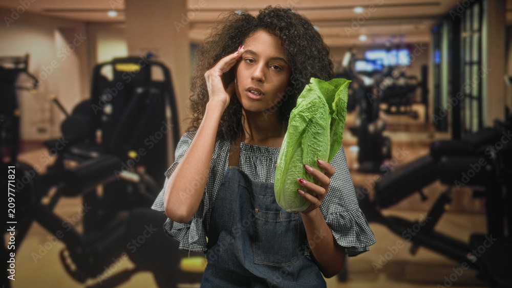 Fototapeta premium Woman in apron holding romaine lettuce and hand to temple for headache inside a gym building, eyes closed and pained; fatigue health concern irony.