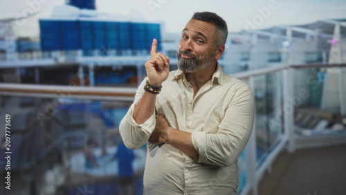Canvas Print Man points index finger upward on cruise ship deck with crossed arms, bearded middle aged hispanic wearing bracelets and casual shirt near railing; confident charm